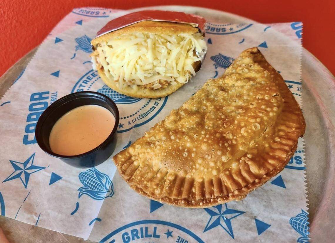 A high-angle shot features a large, golden-brown fried empanada and an arepa overflowing with shredded white cheese. They are served on blue-and-white branded paper from “SABOR STREET GRILL” with a small side of pink dipping sauce.