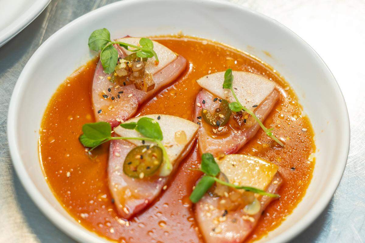 A close-up, high-angle shot of a vibrant seafood dish served in a white bowl. Four triangular slices of raw fish, likely tuna or a similar white fish, with a reddish-pink edge, are arranged in a shallow pool of bright orange-red sauce. Each slice is topped with a small dollop of a relish-like mixture, a thin slice of green jalapeño, and garnished with fresh green microgreens. Black and white sesame seeds are sprinkled throughout the sauce and on the fish. The bowl sits on a metallic surface, possibly a kitchen counter.