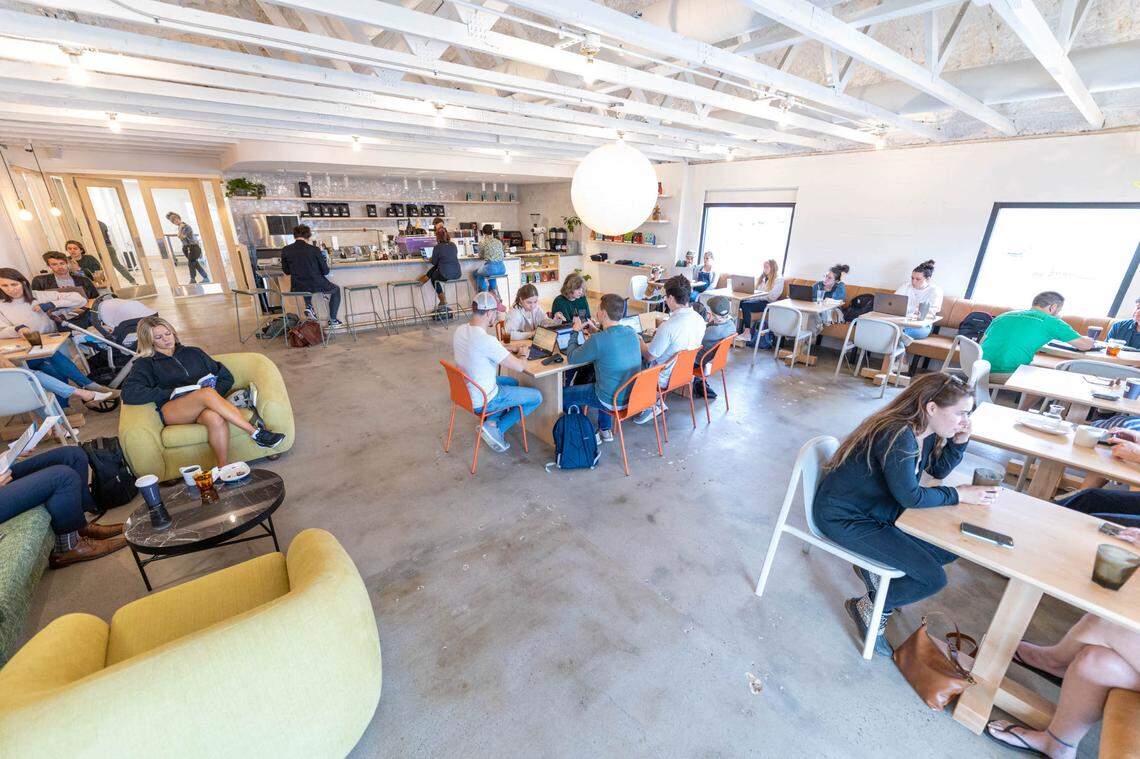 The interior of Night Swim Coffee, a bright, spacious, and modern coffee shop with a high white ceiling exposing beams. The floor is polished concrete. Customers are seated at various tables and on soft seating, many using laptops. In the background, there is a long bar/counter with a few people seated on stools. The furniture includes light wood tables, white and orange chairs, and a few oversized chartreuse and moss-green lounge chairs. A large, spherical white pendant light hangs from the ceiling over a central communal table.