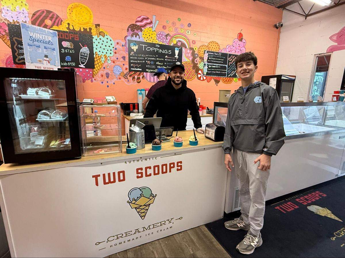 A shop employee and teenager pose behind a white “Two Scoops Creamery” counter. Four cups of ice cream are lined up on the counter between them. The background features the signature orange mural and topping menus.