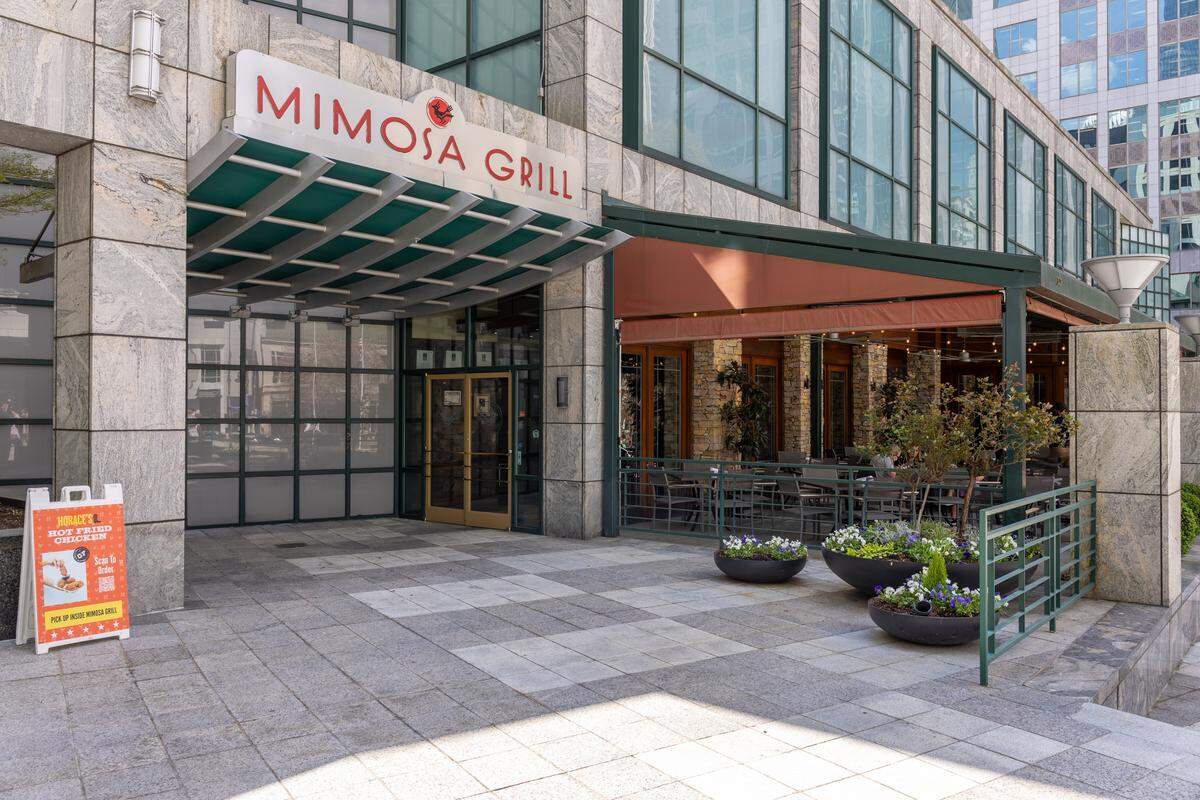 An architectural shot of the Mimosa Grill exterior. The entrance has a large “Mimosa Grill” sign over a green tiered awning. To the right, an outdoor patio is partially covered by a red awning and surrounded by a green metal railing and large potted plants.