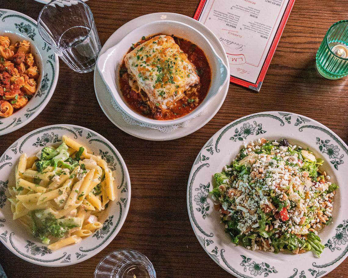 A bird’s-eye view of a wooden restaurant table featuring four Italian dishes: a baked lasagna in a red sauce, penne with broccoli in a cream sauce, a pasta with meat sauce, and a large chopped salad topped with cheese.