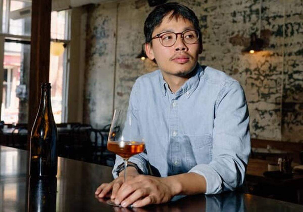A portrait of a chef with glasses and a light blue button-down shirt sitting at a dark, polished bar. The chef is looking off-camera with a neutral expression, resting hands near a glass of amber-colored liquid. Behind the chef, a dark bottle sits on the bar, and the background features a rustic, distressed white brick wall and dim lighting.