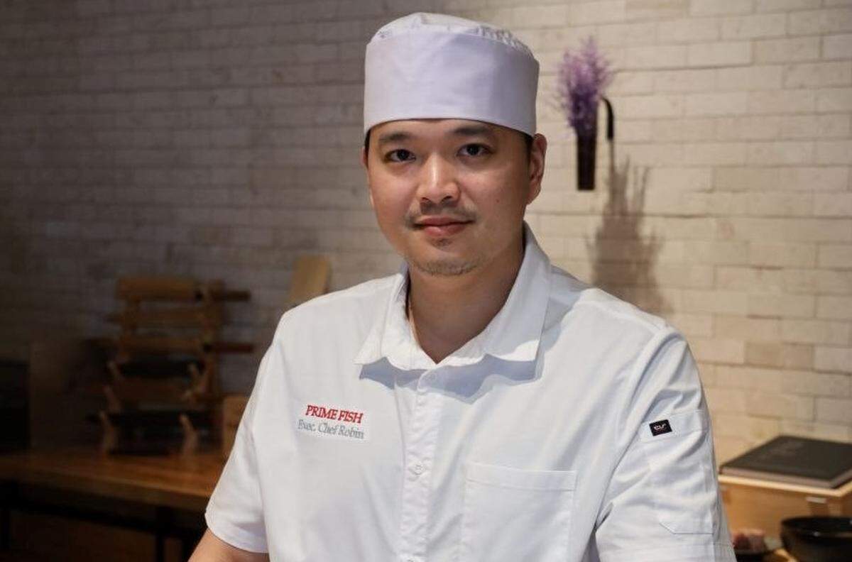 A portrait of a chef wearing a white chef’s coat embroidered with “PRIME FISH Exec. Chef Robin” and a tall white chef’s hat. The chef is standing in front of a light-colored brick wall with a small purple floral arrangement hanging behind. In the background, a wooden counter with a knife rack and other kitchen tools is visible.