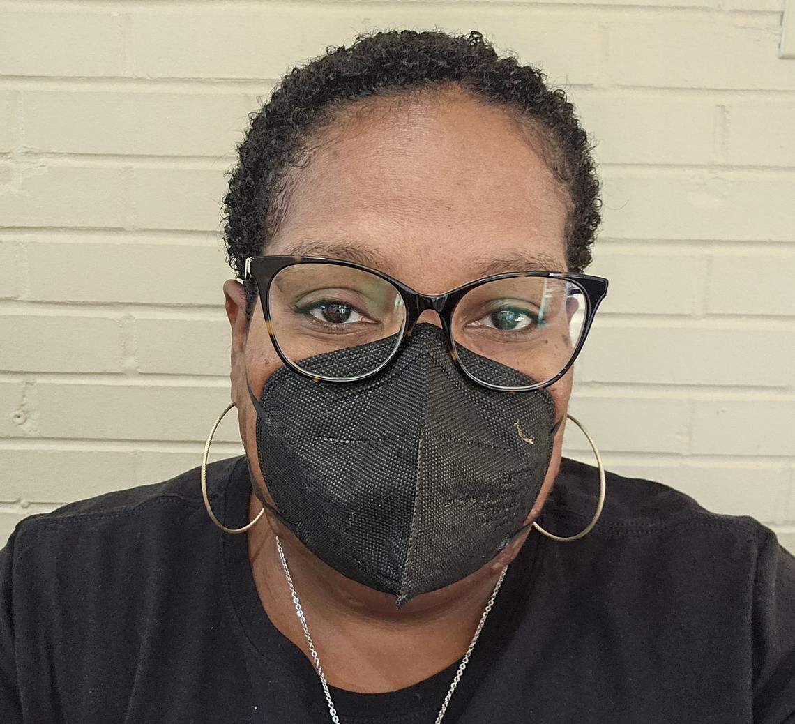A close-up headshot of a person with short, dark, curly hair wearing black-rimmed glasses, large gold hoop earrings, and a black KN95-style face mask. They are looking directly at the camera.
