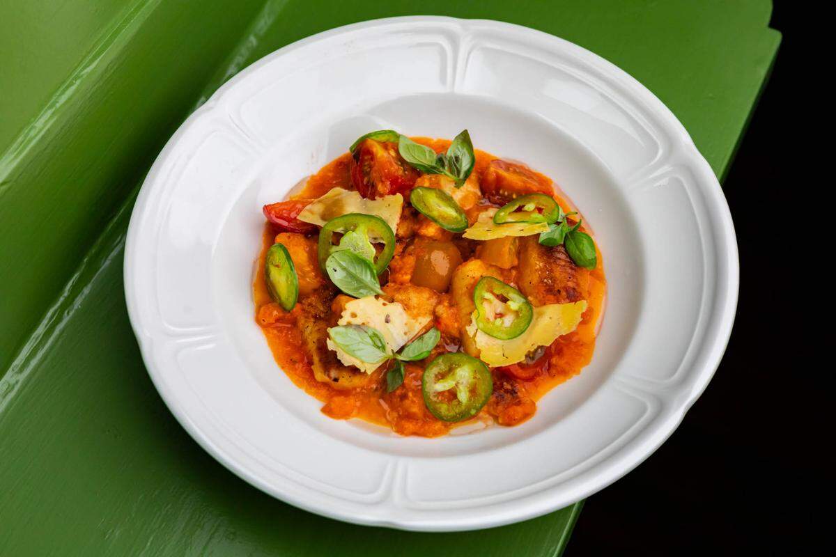 An overhead close-up shot of a white plate holding a dish of ricotta gnocchi arrabbiata, served in a wide, shallow bowl-like plate with a ruffled rim. The gnocchi is tossed in a bright orange-red tomato sauce and garnished generously with slices of green jalapeño pepper, fresh basil leaves, and thin shavings of yellow cheese, likely parmesan. The plate is resting on a glossy, bright green surface, with a black background visible to the right.