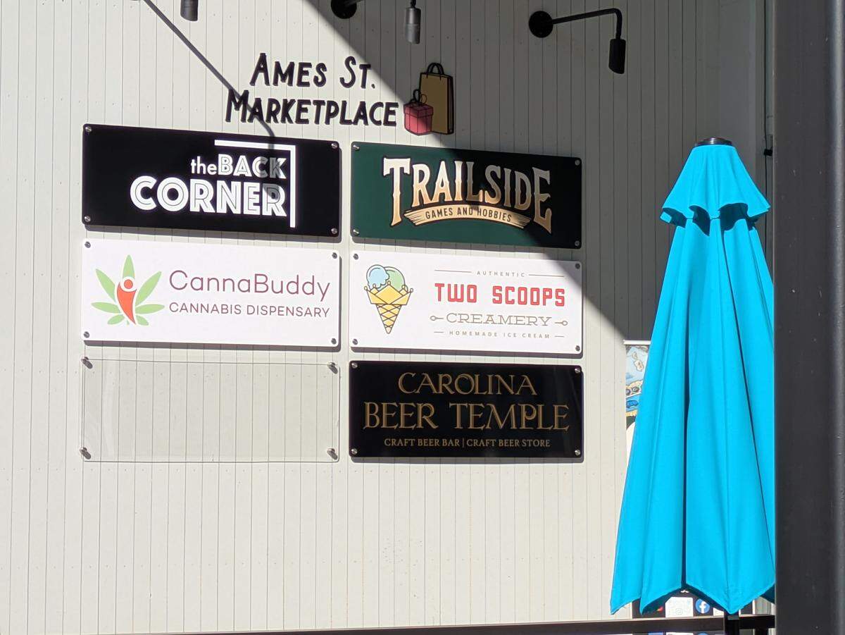 A white exterior wall featuring a directory of rectangular signs for businesses within the Ames St. Marketplace. Visible logos include The Back Corner, Trailside Games and Hobbies, CannaBuddy Cannabis Dispensary, Two Scoops Creamery, and Carolina Beer Temple.
