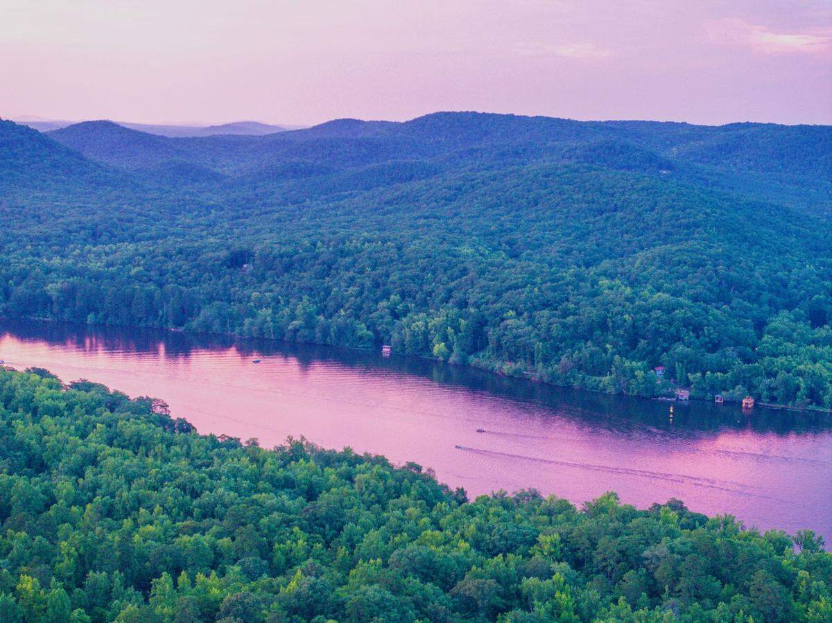 An aerial view of a river winding through the densely forested hills of the Uwharrie National Forest at sunset, with the scene cast in a vibrant pink and purple light.