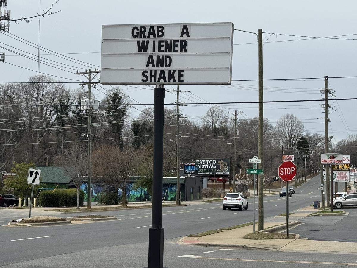 A tall, white roadside marquee sign that reads “GRAB A WIENER AND SHAKE” in bold black letters. The sign is positioned next to a street with cars and utility poles under an overcast sky.