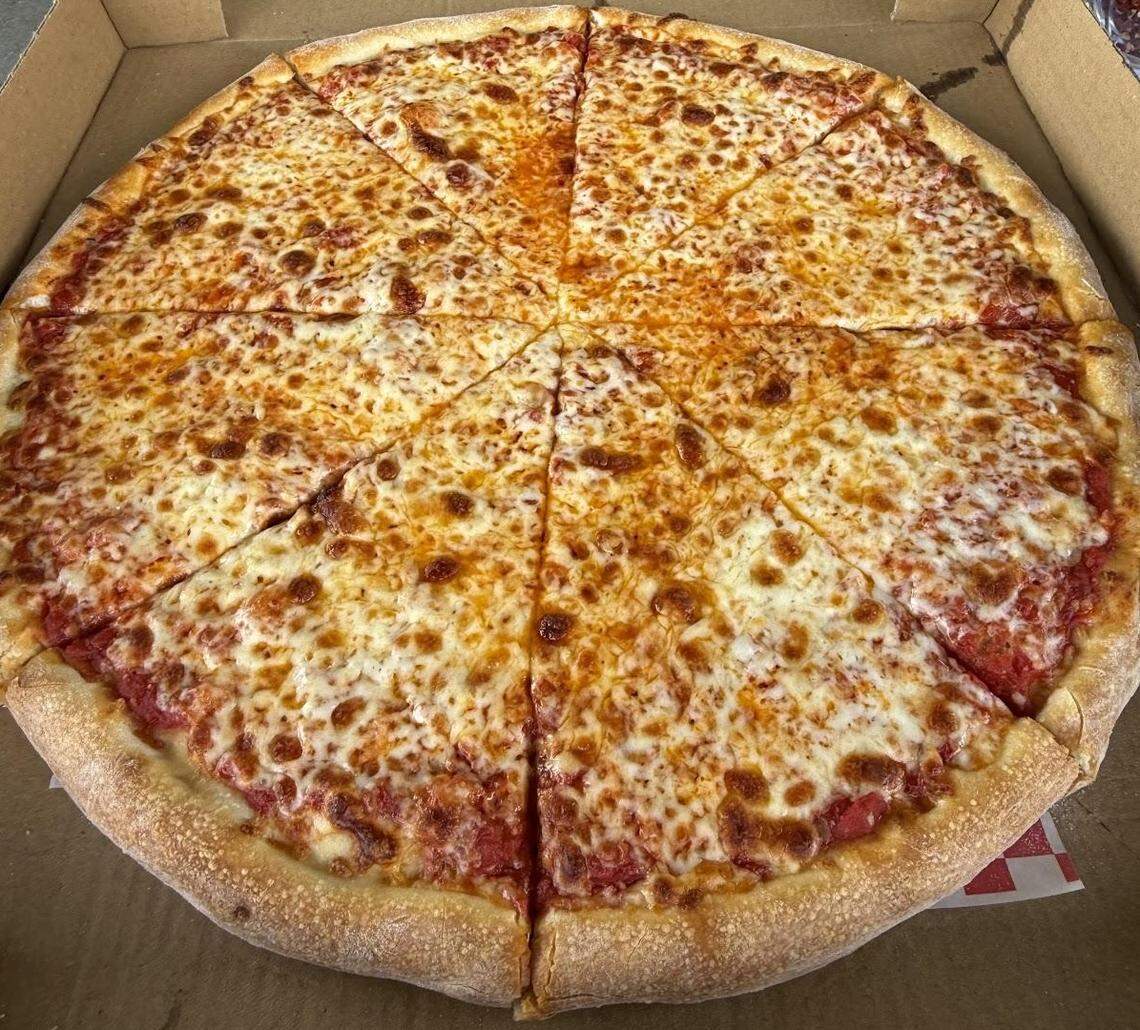 A top-down shot of a large, round cheese pizza in a cardboard takeout box. The pizza is cut into eight slices, with a generous layer of melted, bubbly cheese with dark brown spots. The red sauce is visible in some areas. The crust is thick and golden-brown.