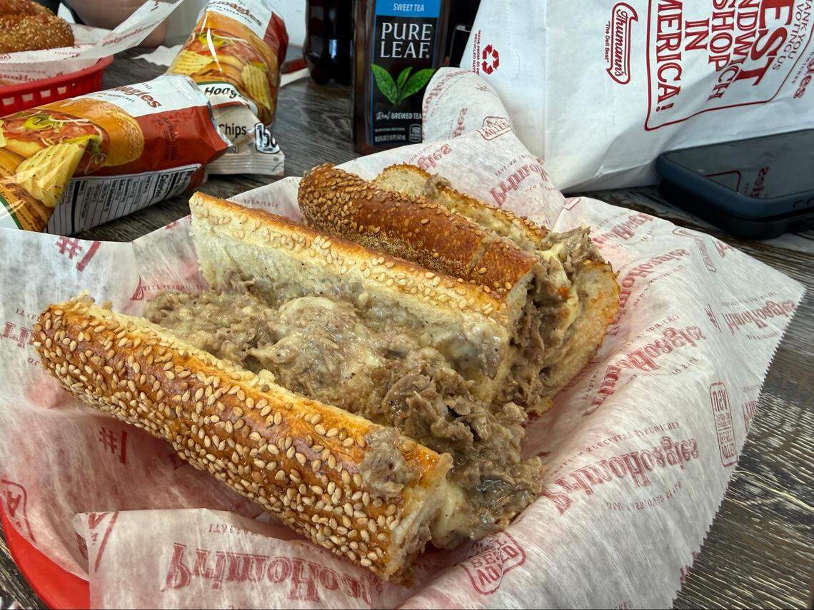 A close-up of a large cheesesteak hoagie sliced into sections and served in a red basket. The sandwich is packed with chopped steak and melted white cheese on a sesame seed roll. Bags of chips and a bottle of Pure Leaf Sweet Tea are in the blurred background.