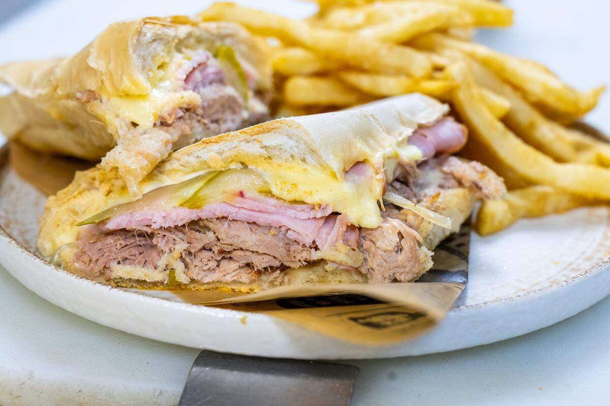 A close-up shot of a pressed Cuban sandwich, cut in half, on a light-colored plate. The cross-section clearly shows layers of shredded pork, sliced ham, melted white cheese, and pickles. A large pile of golden french fries is served alongside it in the background.