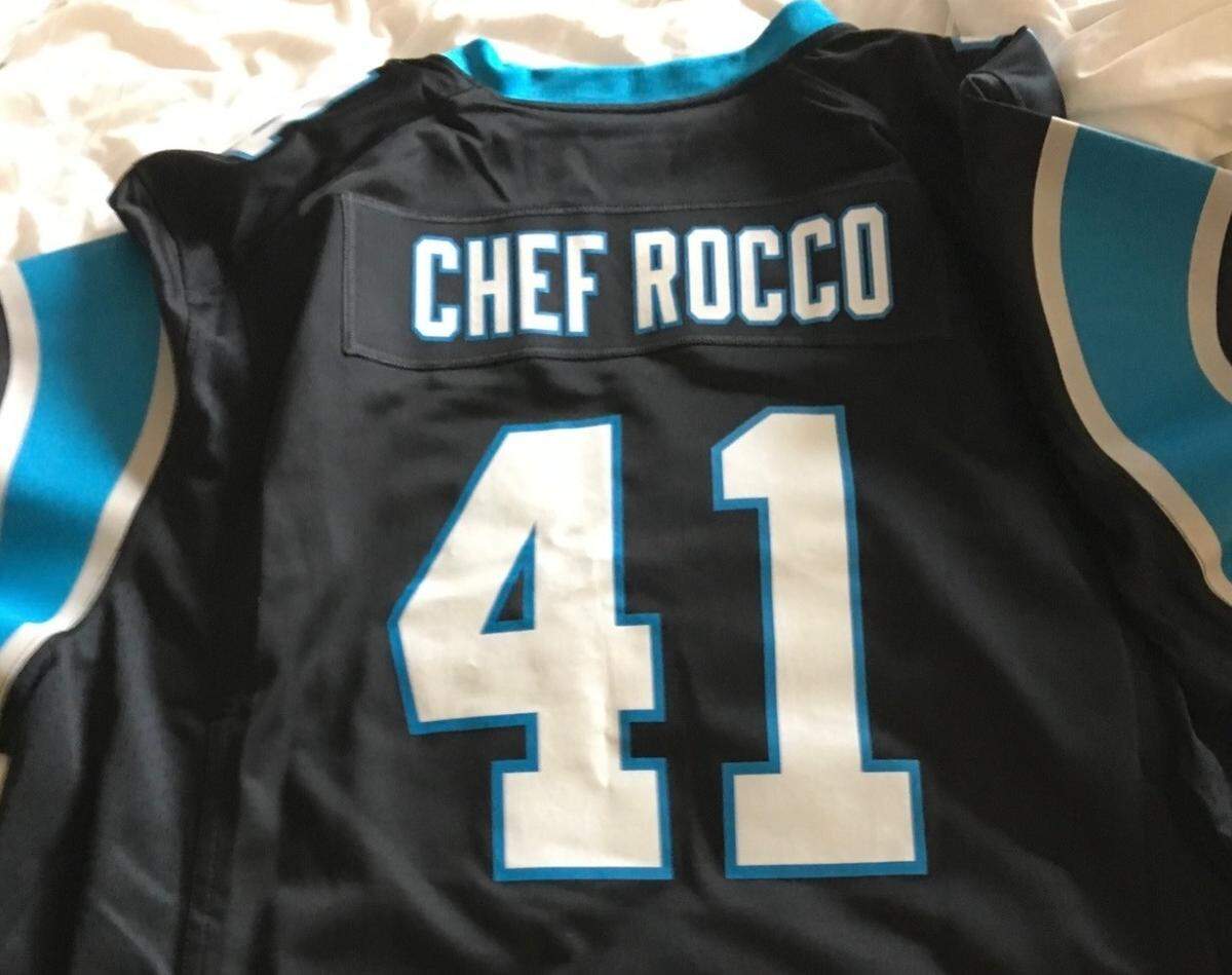 A close-up, high-angle shot of the back of a black Carolina Panthers jersey, lying flat on a light-colored surface. The jersey has light blue trim and features the name “CHEF ROCCO” in white and light blue letters across the top, above the large number “41.” The name and number are stitched or printed onto the jersey.