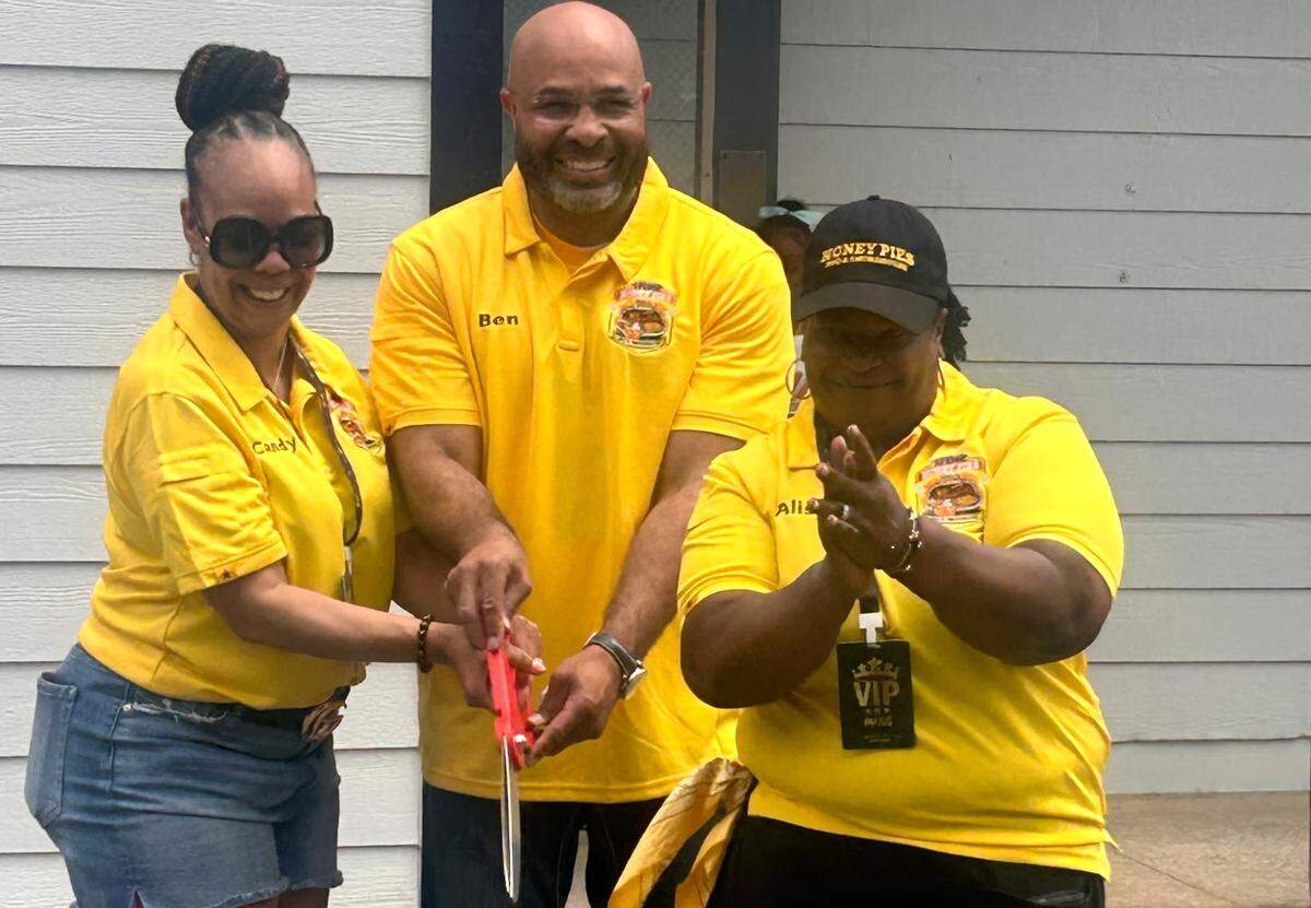 Three people wearing matching bright yellow polo shirts participate in a ribbon-cutting ceremony. The person in the center, whose shirt is embroidered with “Ben,” holds large red ceremonial scissors with the person to his left. A third person on the right claps in celebration. All three shirts feature a logo for “Honey Pies BBQ & Seafood.”