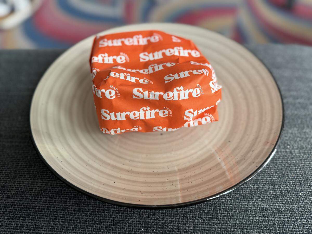A close-up of a sandwich tightly wrapped in signature orange paper with a repeating white “Surefire Market” logo. It sits on a beige ceramic plate with a subtle concentric ring pattern