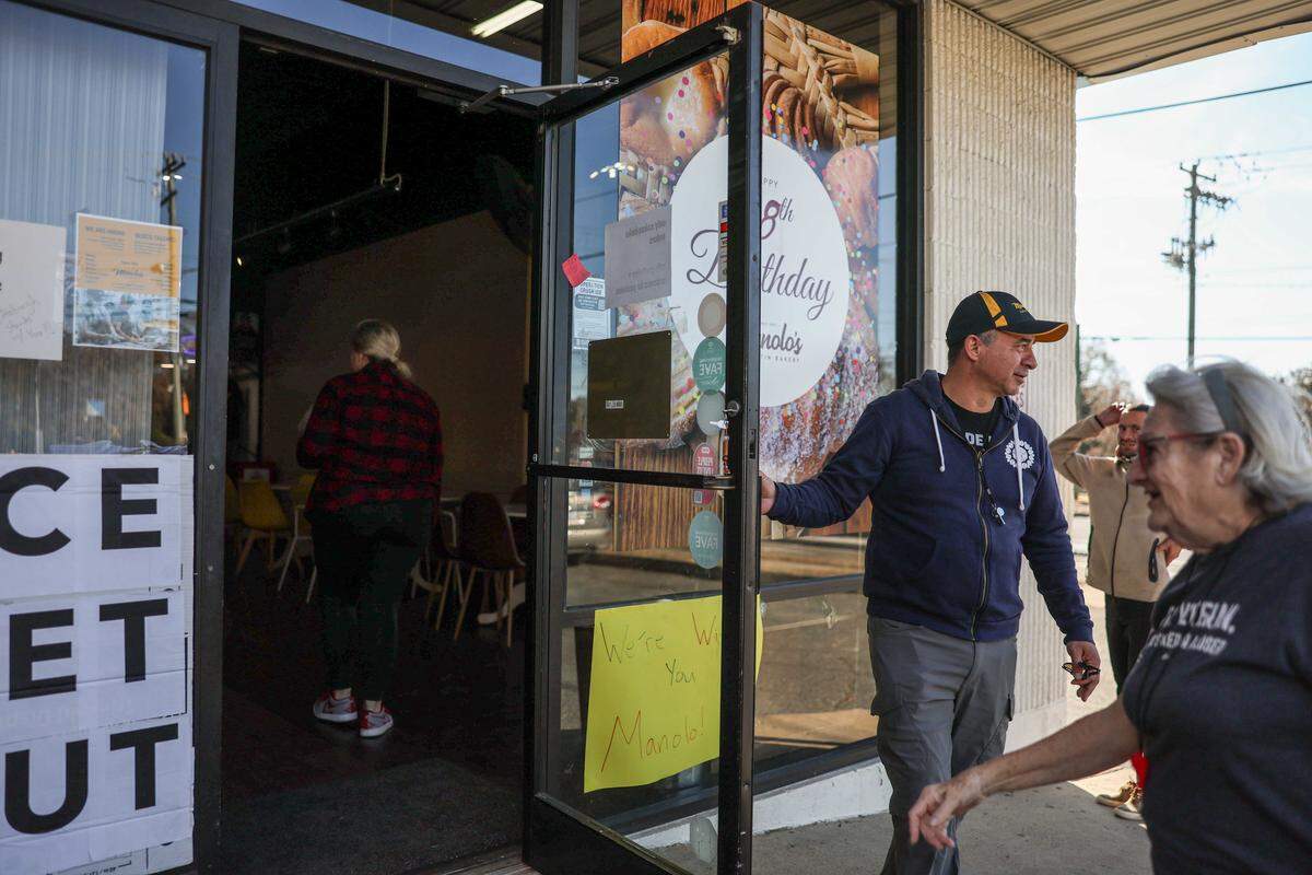 An eye-level shot of the open glass entrance door of a bakery. The owner, in a navy hoodie, holds the door for a person walking past. The door is covered with multiple signs, prominently featuring a yellow note that reads, “We’re With You Manolo!”