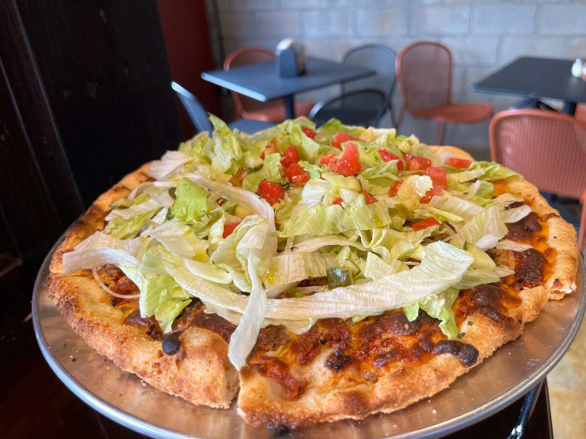A vibrant, fully loaded bacon cheeseburger pizza sits on a metal pizza pan in what appears to be a casual restaurant setting. The pizza has a golden-brown, crispy crust and is generously topped with a layer of melted cheese and what looks like seasoned ground meat. A large mound of fresh, shredded iceberg lettuce covers the entire pizza, and it’s generously sprinkled with diced red tomatoes and what appear to be pickled jalapeños or cucumbers. In the blurred background, several dark tables with chairs are visible against a light-colored brick wall.