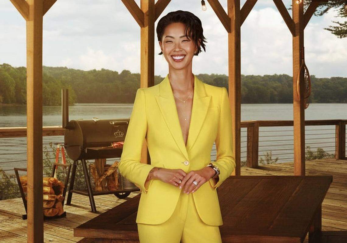 A person is standing at the center of the frame with a bright smile, wearing a vibrant yellow power suit with a single-button blazer and matching trousers, accented by a gold lariat necklace and a silver watch. The photo is positioned on a rustic wooden deck under a pergola with exposed beams.