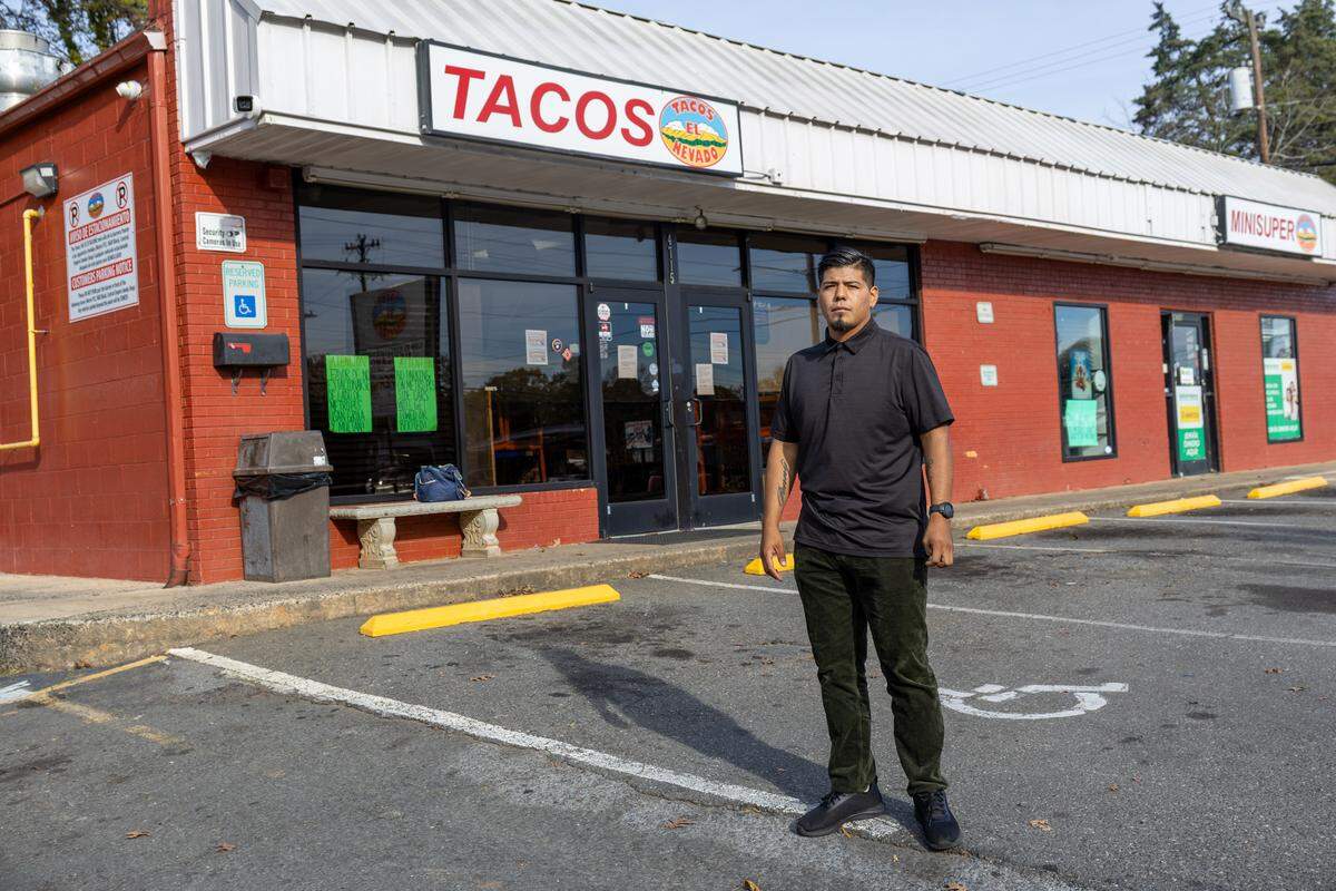 A full-body, eye-level shot of a restaurant worker wearing a black polo shirt and dark pants, standing in an asphalt parking lot. The worker is positioned in front of the red brick exterior of a business whose main sign reads “TACOS” in large red letters. A “MINISUPER” sign is also visible on the right side of the building.