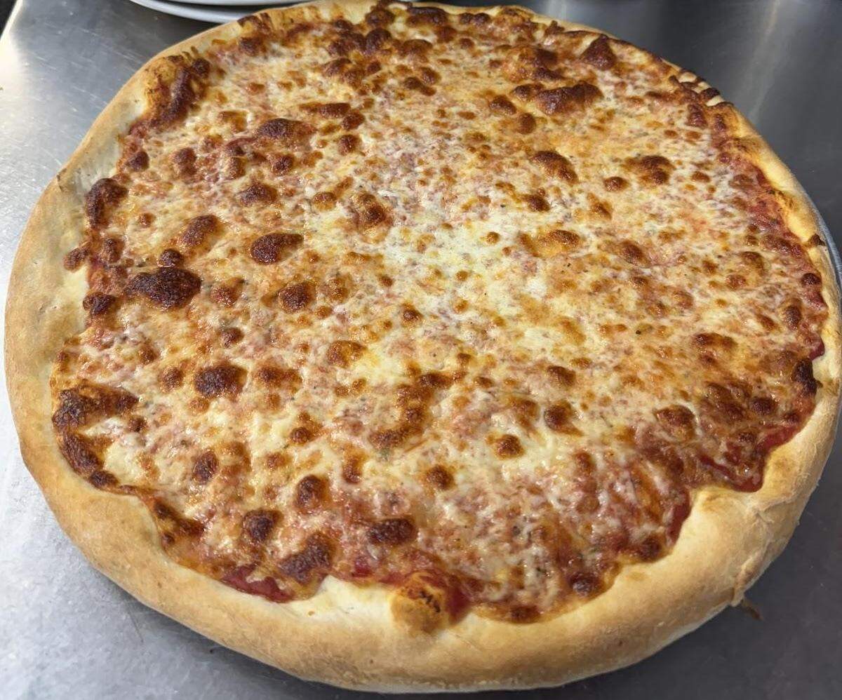 A high-angle, close-up shot of a classic round cheese pizza, fresh out of the oven. The pizza has a thick, golden-brown crust and is covered with a melted, bubbly layer of cheese with dark, caramelized spots. The red tomato sauce is visible in some areas underneath the cheese. The pizza is sitting on a metallic, possibly stainless steel, surface.