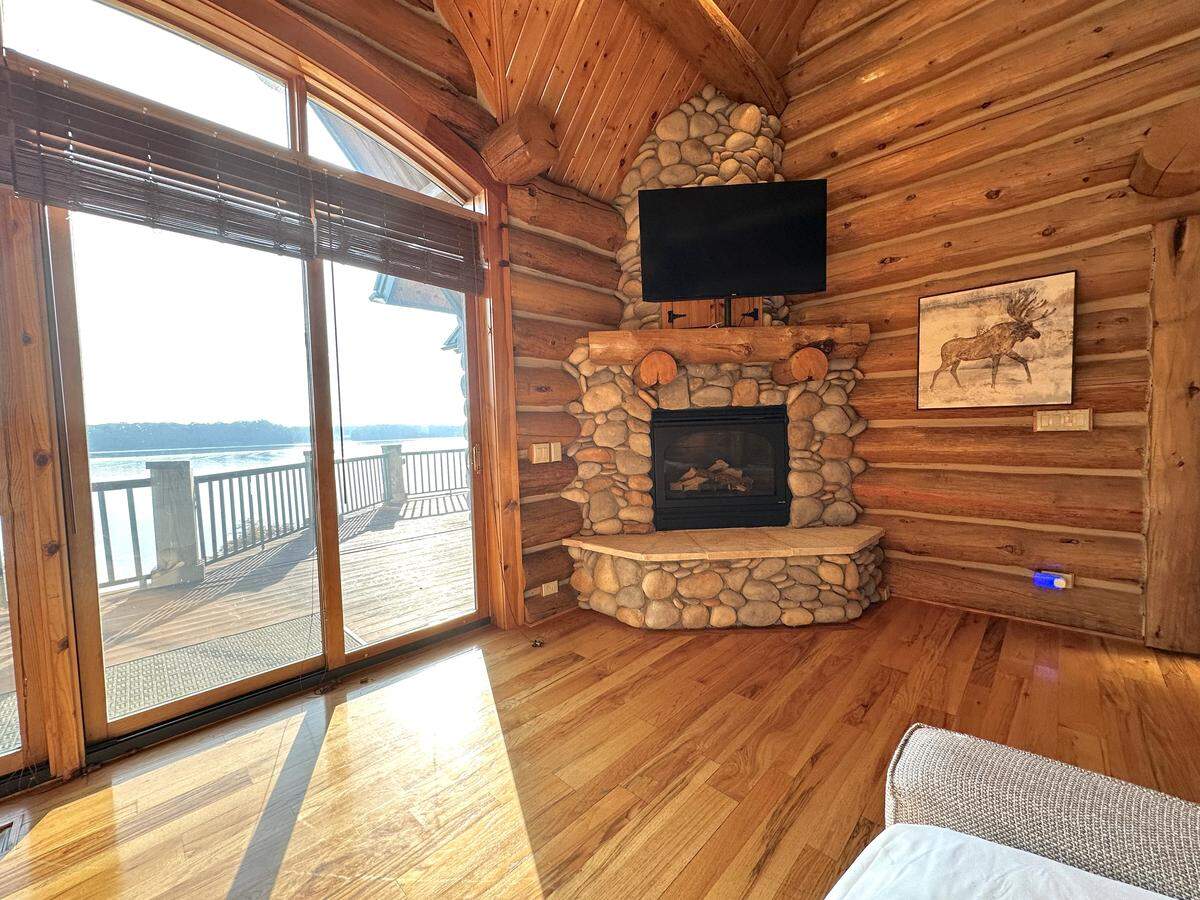 A sitting area in a log cabin bedroom, featuring a corner fireplace made of river stones with a TV mounted above it. Large glass doors to the left of the fireplace lead to a deck overlooking a sunny lake. A painting of a moose hangs on the log wall.