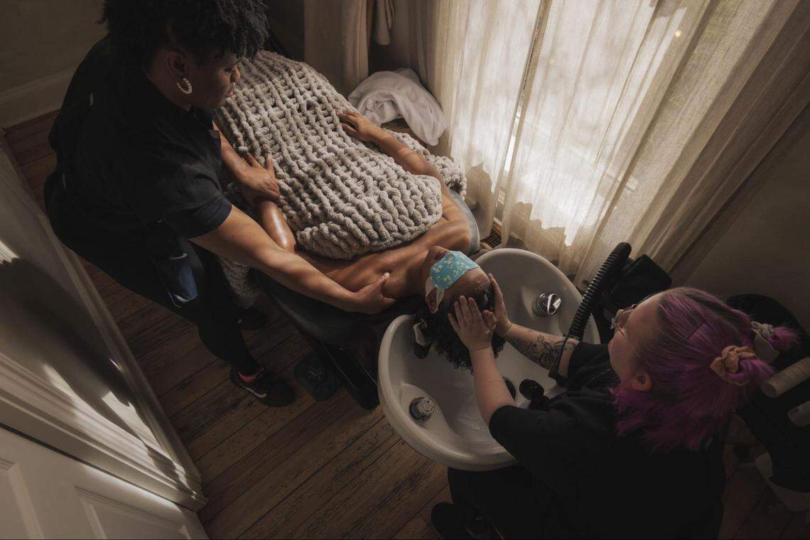An overhead view of a client receiving a dual treatment. One technician massages the client’s shoulders while another washes their hair in a white basin. The client is covered with a thick, chunky knit blanket.