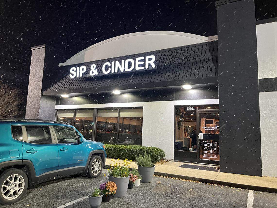 An eye-level, wide shot of the “SIP & CINDER” restaurant exterior at night during a light snowfall. A teal SUV is parked in the foreground on the left, and the building, featuring a lit sign and large windows, is on the right, with potted plants by the entrance.