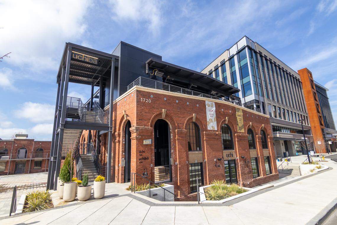 An eye-level, wide-angle shot shows the exterior of a multi-story building that blends historic and modern architecture. The foreground features a two-story red brick building with large, prominent arched windows and stone accents.