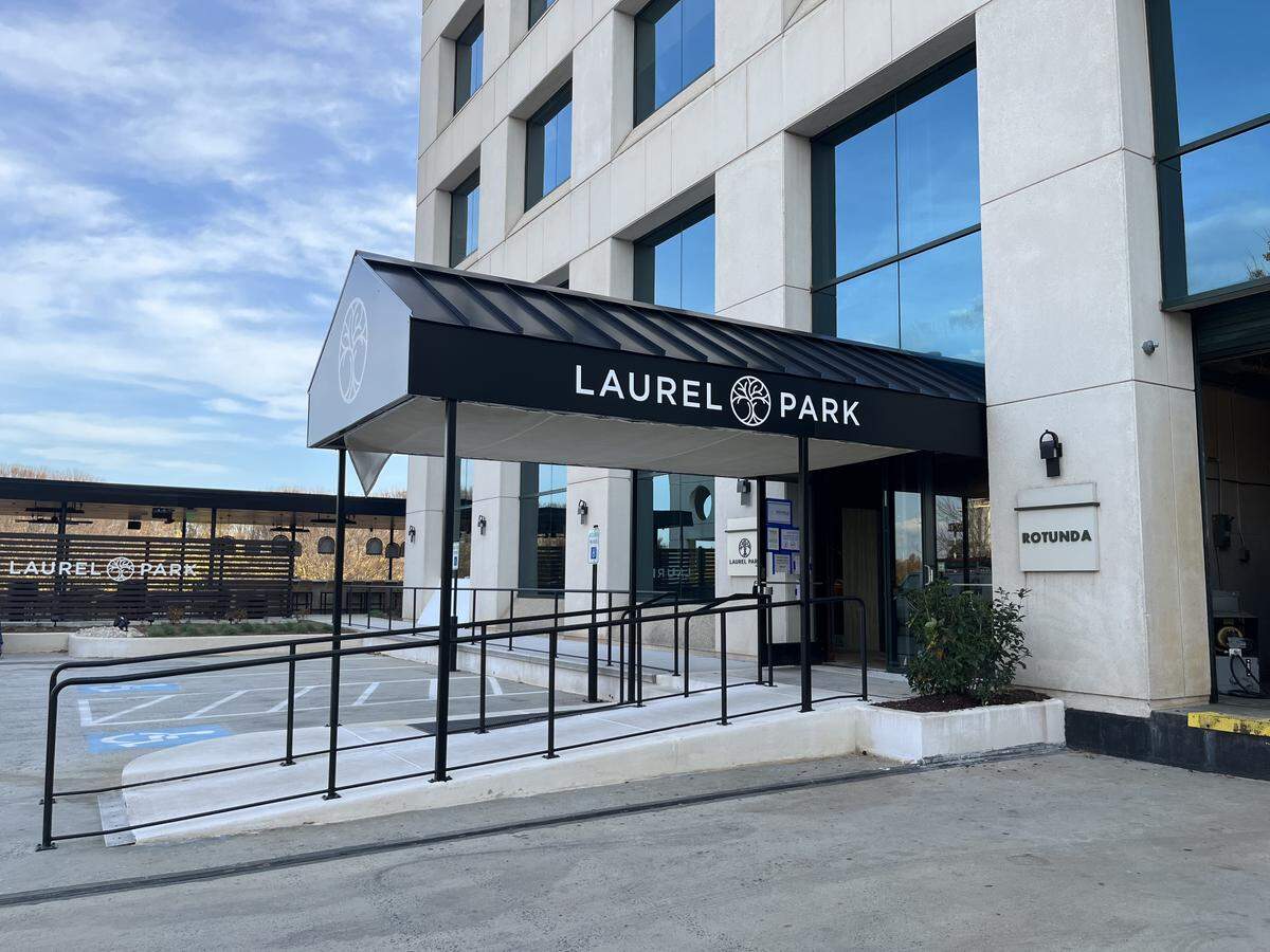 An eye-level, angled shot of the main entrance of the multi-story Laurel Park building. A black awning with the white “LAUREL PARK” text and logo covers the entrance, which is accessed by a long, black-railed accessibility ramp. The building has a light-colored stone facade and large reflective windows.