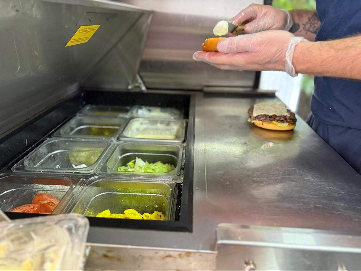 A close-up, high-angle shot of a person’s hands wearing clear plastic gloves, preparing a burger on a food truck’s stainless steel countertop. The person is holding a bun with a piece of cheese on it. On the counter, a finished burger patty with melted cheese and pickles is visible on the bottom bun. A built-in cooler on the counter holds various ingredients in clear plastic bins, including sliced tomatoes, lettuce, and pickles.