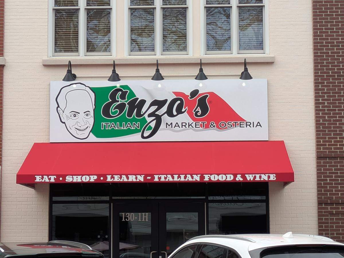 The storefront of “Enzo’s Italian Market & Osteria” featuring a large sign with the Italian flag colors and a caricature of a man’s face. A red awning over the entrance says “Eat • Shop • Learn - Italian Food & Wine.”