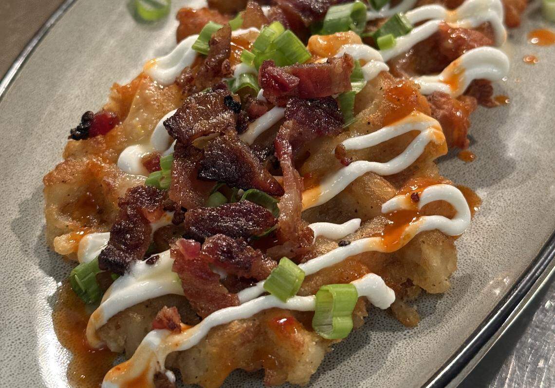 A close-up, top-down shot of a savory waffle appetizer served on a gray ceramic plate. The golden-brown waffle is loaded with toppings, including crispy bacon bits, chopped green onions, a zigzag drizzle of white crema or sour cream, and a spicy orange buffalo-style sauce. The lighting is warm and bright, highlighting the textures of the crispy toppings and the saucy finish.