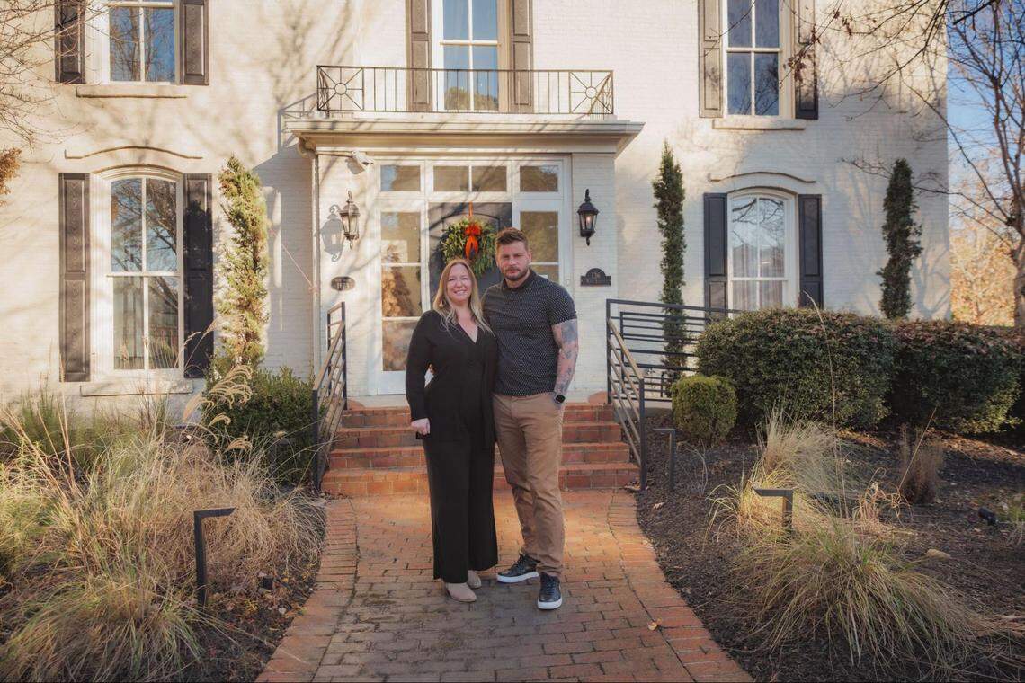 A couple stand together in front of a large, two-story white brick house with black shutters. They are dressed in casual attire, and the home features a brick walkway, manicured landscaping, and a wreath on the front door.