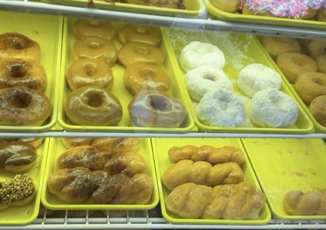 Looking into a glass display case at a donut shop, multiple yellow trays hold different types of donuts. On the top shelf are maple bars and powdered sugar donuts. On the bottom shelf are rows of glazed and plain twist donuts.