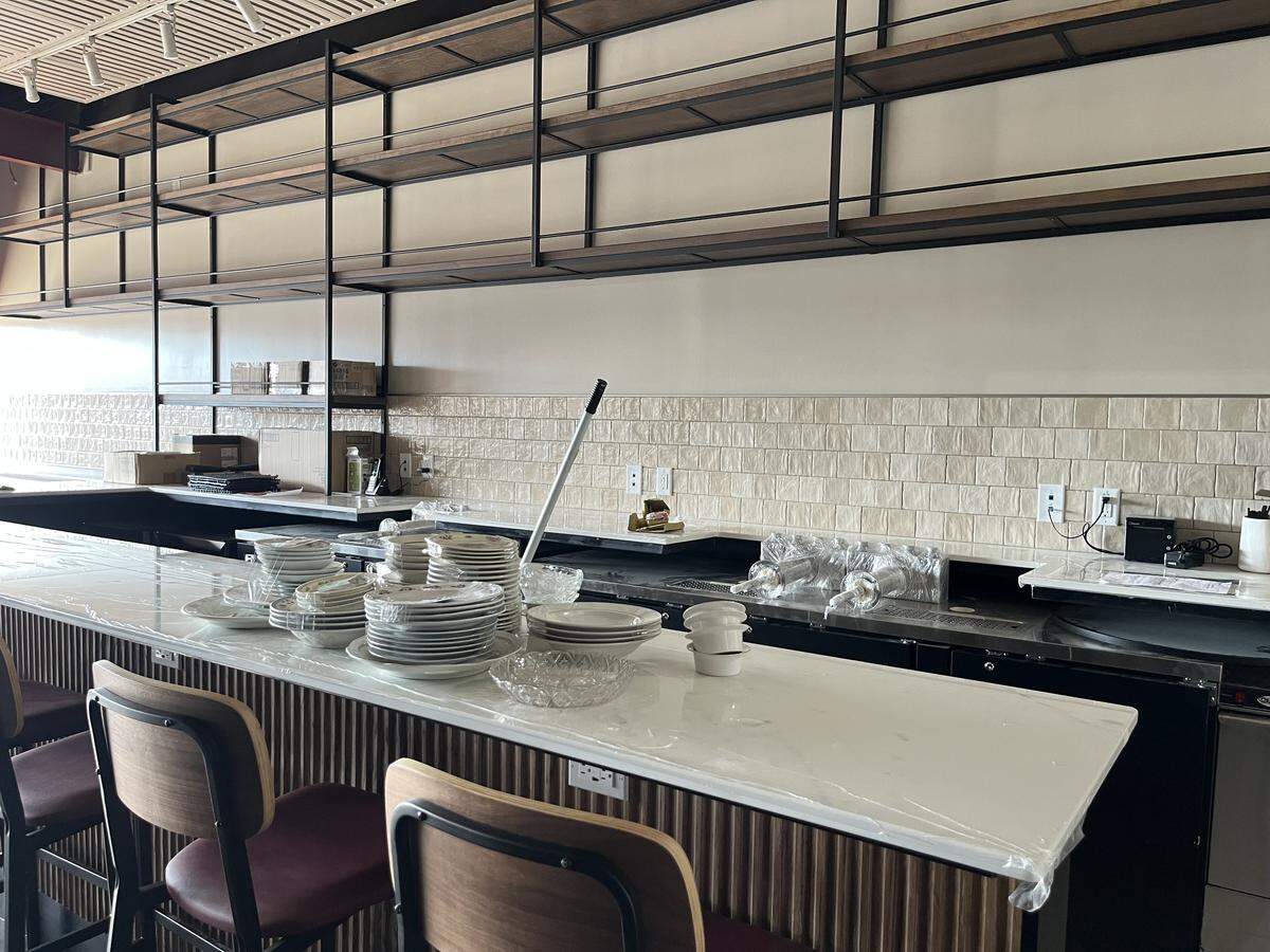 The interior of a bar or restaurant under construction or being set up. The foreground features a long white countertop, partially covered in plastic sheeting, with stacks of clean white plates and bowls on it. Two bar stools with dark red upholstered seats and wood backs are visible. Behind the bar, there is a backsplash of subway tile and a back bar shelving unit with open dark wood and black metal shelves. The space appears to be newly renovated or prepared for opening.
