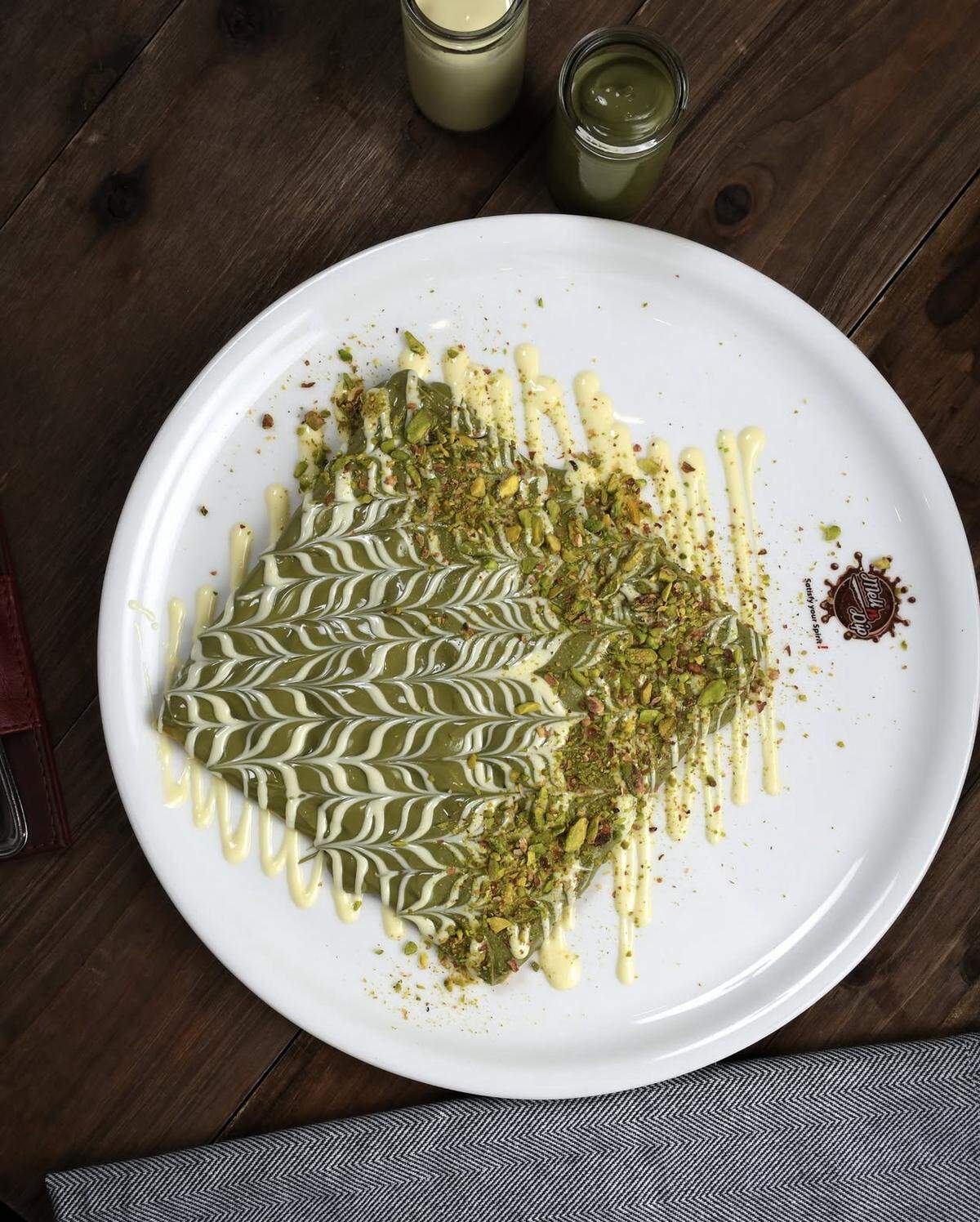 A top-down view of a large dessert crepe served on a round white plate. The crepe is completely covered in a vibrant green pistachio sauce and intricately drizzled with a white chocolate pattern. One half is further topped with a generous layer of crushed pistachios. Two small glass jars containing additional green and white sauces are visible in the background on a dark wooden table.