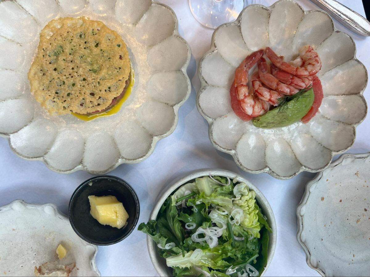 A top-down shot of a table with several small plates of food, likely from a shared meal. On the left, a scalloped, off-white ceramic plate holds a yellow and green tuna tartare topped with a large, delicate, lacy cheese crisp. On the right, a matching scalloped plate holds several pink shrimp and a scoop of mashed avocado. In the foreground, a small white bowl contains a green salad with thinly sliced white onions. A small black dish with a piece of yellow butter or cheese is also visible. The plates are set on a white tablecloth.
