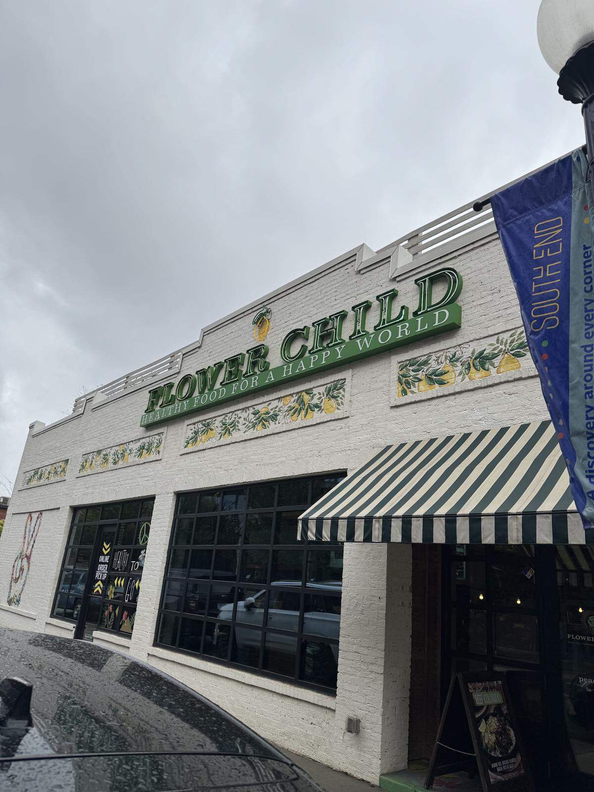 Exterior view of a restaurant named Flower Child. The sign is green and reads, “HEALTHY FOOD FOR A HAPPY WORLD” underneath. The building has a white brick facade with lemon-themed tiles and a green and white striped awning over the entrance.