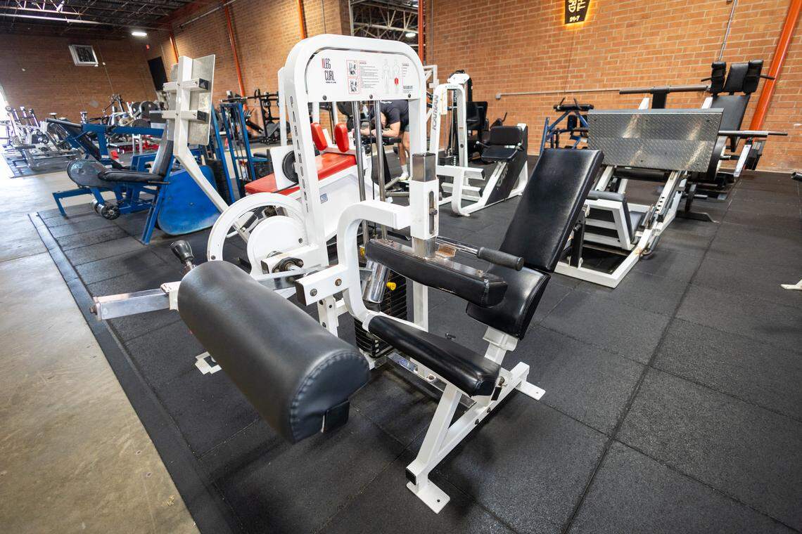 A white leg curl machine with black padding sits on a black gym mat. The machine has exposed cables and a weight stack visible in the center.
