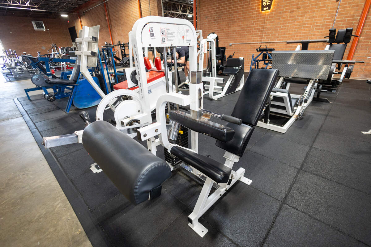 A white leg curl machine with black padding sits on a black gym mat. The machine has exposed cables and a weight stack visible in the center.