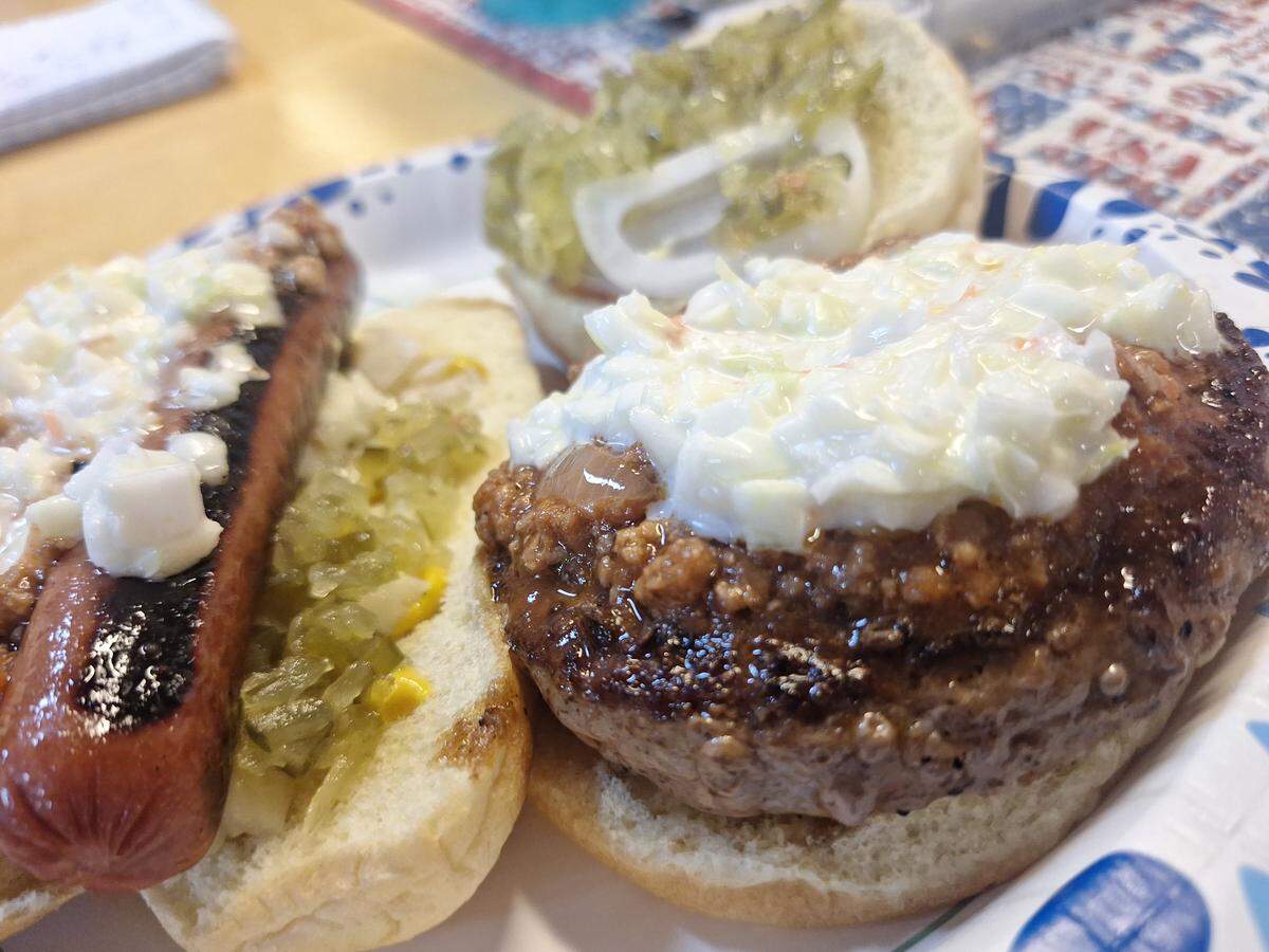 A close-up shot of a paper plate holding a hot dog and a hamburger, both prepared in a classic American style. On the left, a hot dog in a toasted bun is topped with pickle relish and what appears to be a creamy coleslaw. On the right, a large burger patty sits on a bun, topped with coleslaw. Another open bun in the background holds a relish topping and white onion slices. The plate is on a wooden table, with a colorful placemat visible underneath.