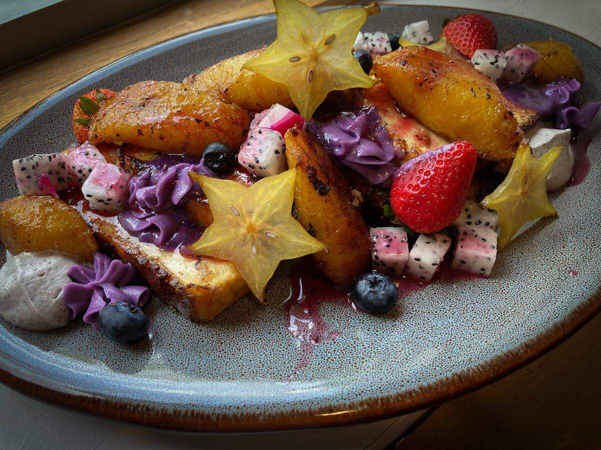 A beautifully composed brunch dish of French toast bake on an oval plate. The dish is a riot of color, decorated with translucent, star-shaped slices of starfruit, cubes of white dragon fruit, blueberries, strawberries, and caramelized plantains, all interspersed with piped dollops of a rich purple cream.