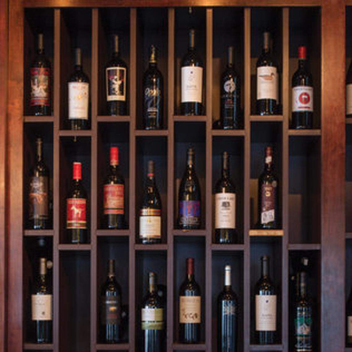 A grid of dark wood shelving holds an assortment of approximately twenty-four wine bottles, each with different labels and capsule colors. The display is mounted on a wall and features red wines and dark bottles arranged symmetrically across three vertical columns and eight horizontal rows.