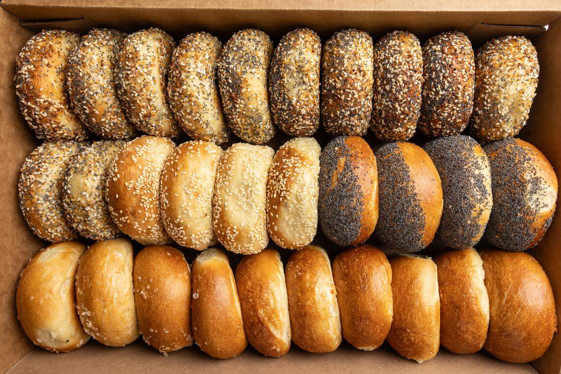 A full, overhead shot of a Classic Bagel Box showing two dozen bagels neatly arranged in three rows inside a cardboard box. The bagels feature a variety of toppings, including everything, poppy seeds, sesame seeds, and plain.