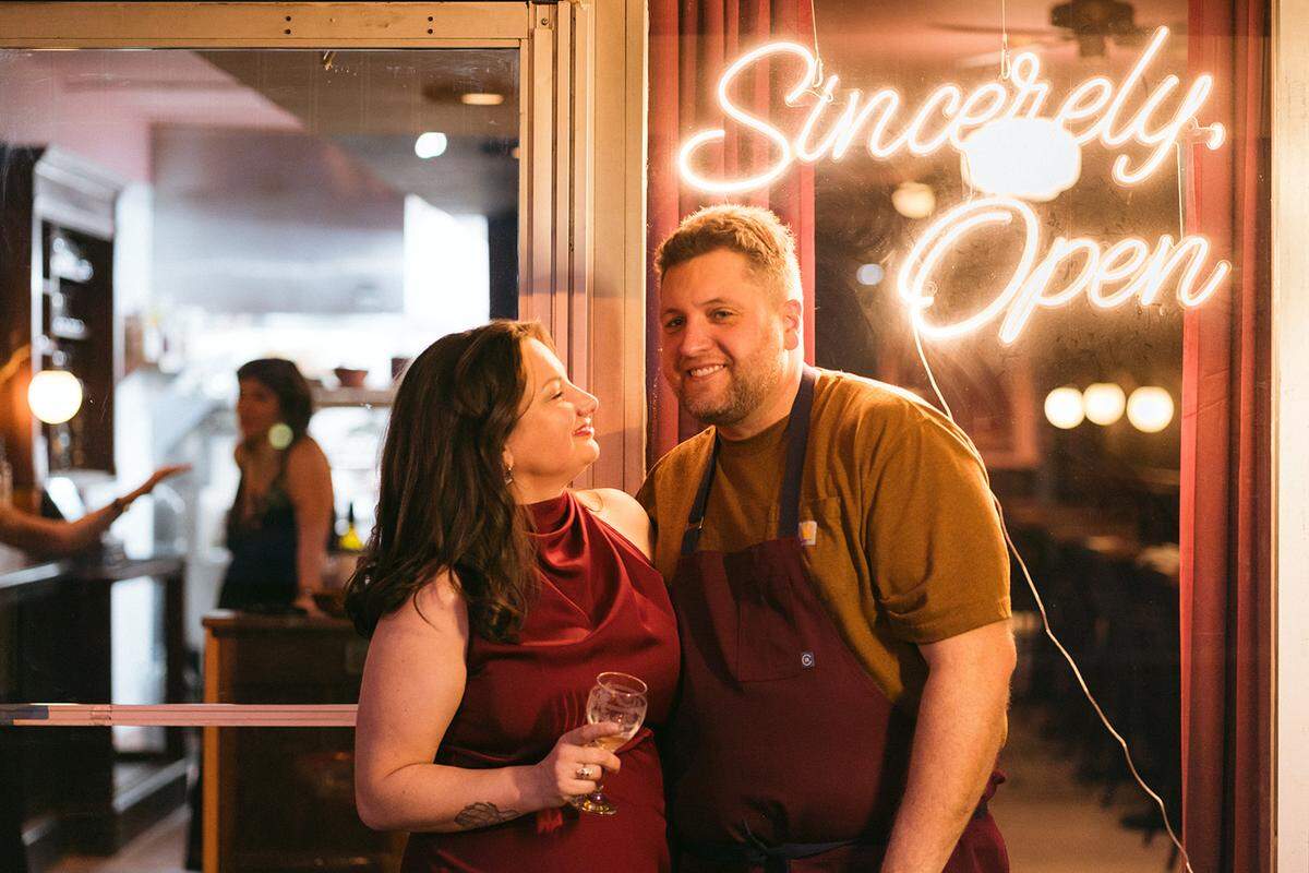 A warm, candid portrait of a woman and a man standing together in front of a window. The woman, on the left, wears a sleeveless burgundy dress and holds a wine glass, looking up at the man with a smile. The man, on the right, wears a mustard-colored shirt and a dark burgundy apron, smiling toward the camera. Above them, a glowing neon sign in the window reads “Sincerely, Open.” The background shows the soft, blurred interior of a restaurant.