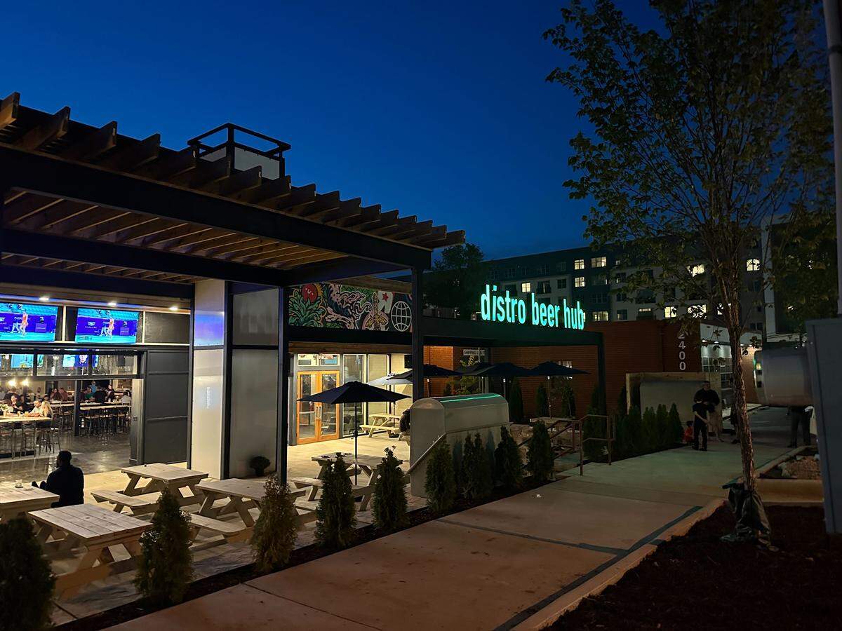 A nighttime shot of the Distro Beer Hub building, with its name illuminated in green neon letters. The patio is visible with picnic tables and a few people enjoying the evening under the dark blue sky.