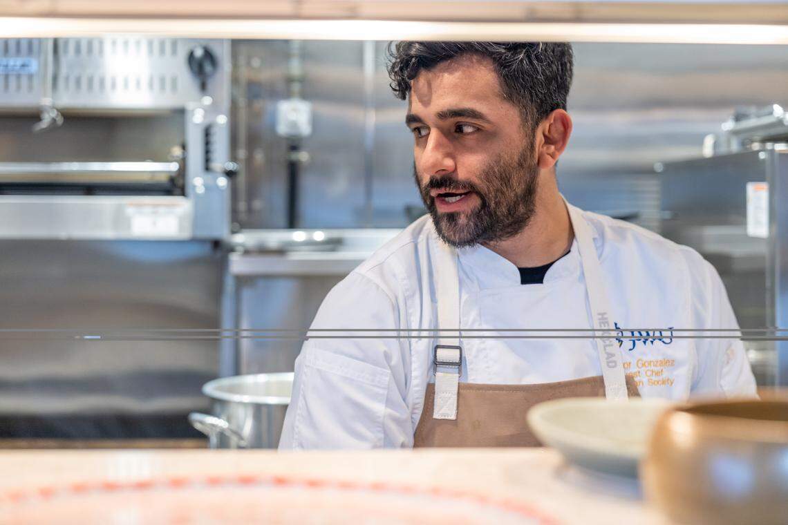 A chef with dark, curly hair and a beard is shown from the chest up, wearing a white chef’s coat and a tan apron branded with “JWU” and looking off-camera to his left with a focused expression while speaking. The shot is taken through a low, horizontal opening, likely a kitchen pass, with blurred plates in the immediate foreground and a professional stainless steel kitchen in the background.