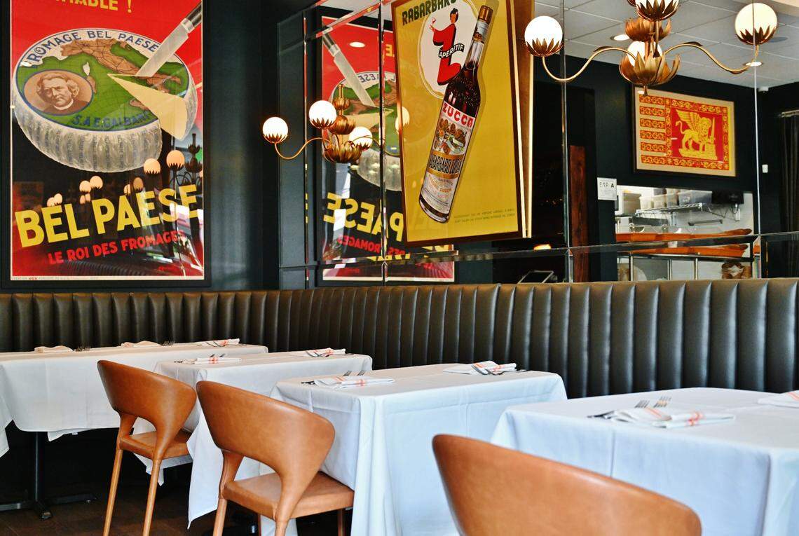 A bustling indoor taproom featuring a long bar with several patrons seated on wooden stools. Behind the bar, a large chalkboard menu displays a variety of beers above a row of silver taps. The space is decorated with orange accent walls, hanging track lighting, and a dark wooden table in the foreground holding a long planter of green grass.