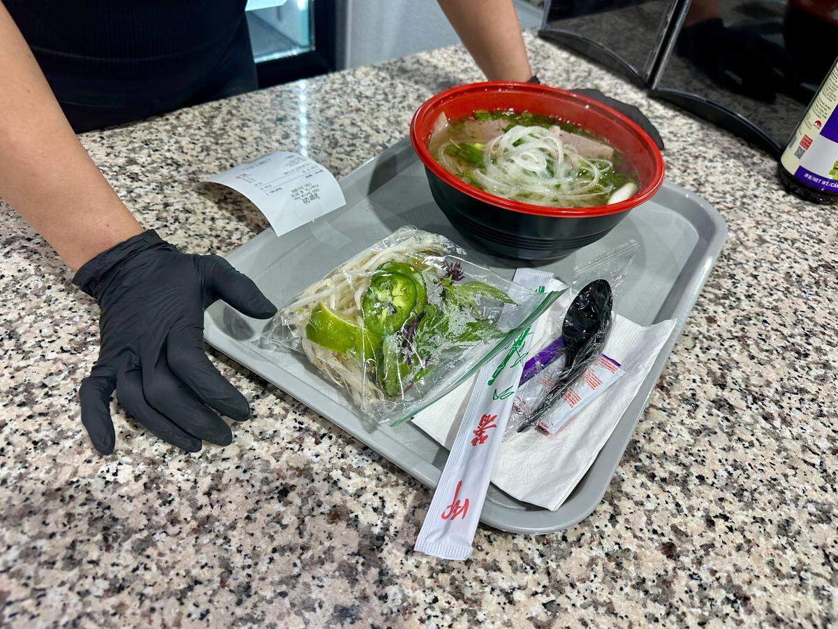 A tray holding a large bowl of pho with a red rim, filled with broth, noodles, and sliced white onions. The tray also includes a small plastic bag of fresh bean sprouts, jalapeños, lime, and herbs, along with wrapped chopsticks and a plastic spoon. A person wearing black gloves is shown placing the tray on a granite countertop next to a printed receipt.