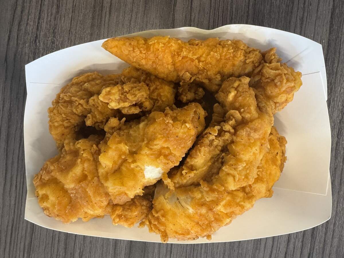 A close-up, top-down shot of several large, thick fried chicken tenders piled inside a small white paper food boat. The chicken strips feature a bright golden-orange, craggy, and crispy breading, with a piece in the front showing the juicy white meat inside where the crust has broken.